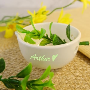 May include: An off-white ceramic bowl with the name "Arthur" and a heart symbol in light green. The bowl contains green leaves and is on a textured surface. Yellow flowers and green leaves are in the background.
