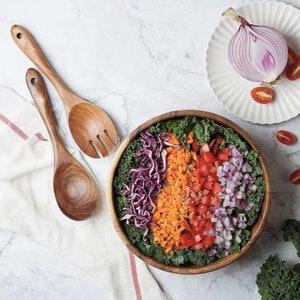 May include: A wooden salad bowl filled with a colorful salad of red cabbage, carrots, tomatoes, red onion, and kale. Two wooden salad serving utensils are on a white marble surface.