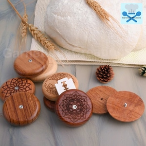 Könnte beinhalten: Ein Satz von sechs Holz-Brotmessern mit komplizierten Designs. Die Messer sind aus Naturholz gefertigt und haben eine scharfe Rasierklinge zum Einschneiden von Brotteig. Die Messer sind auf einer hellbraunen Oberfläche mit einem Brotlaib im Hintergrund zu sehen.