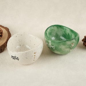 May include: Two decorative bowls. One is speckled white with the name "Arthur" in script. The other is green with a marbled effect and the name "William" in script. Both bowls have an organic shape and are set on a neutral surface.