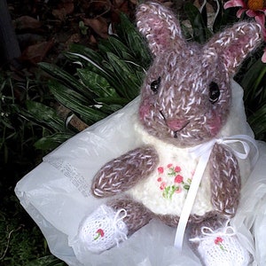May include: A brown and white knitted bunny rabbit wearing a white sweater with pink embroidered flowers. The bunny is sitting in a white plastic bag with a white ribbon around its neck.