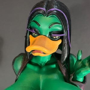 May include: A green figurine of a duck-like character with black hair and purple highlights. The character has an orange beak and is wearing a brown vest. The figurine is a bust, showing the upper body and head.