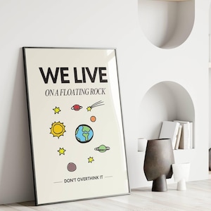 May include: A black framed poster with a white background and the text "WE LIVE ON A FLOATING ROCK - DON'T OVERTHINK IT" in black. The poster features a cartoon illustration of the Earth with planets and stars in a colourful design.