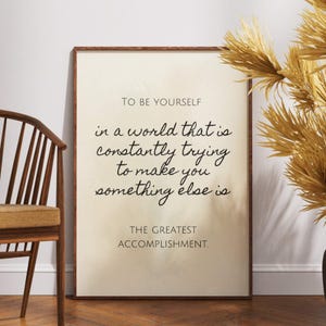 May include: A motivational quote printed on a beige background in a brown frame. The quote reads "To be yourself in a world that is constantly trying to make you something else is the greatest accomplishment."