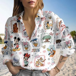 Może przedstawiać: Biała koszula zapinana na guziki z kolorowym wzorem postaci Disneya, zamków i fajerwerków. Koszula zawiera wizerunki Myszki Miki, Myszki Minnie, Kaczora Donalda i Goofy'ego. Rok "2026" jest również nadrukowany.