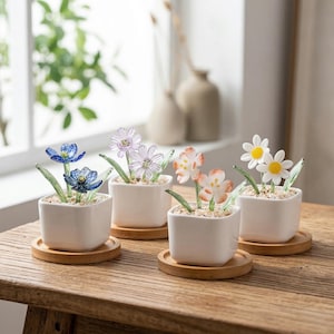 Può includere: Quattro piccoli vasi quadrati in ceramica bianca, ciascuno con una diversa composizione floreale in vetro. I fiori sono blu, lavanda, pesca e bianchi con steli e foglie di vetro verde. Ogni vaso poggia su un piccolo sottobicchiere rotondo in legno.