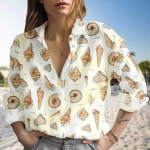 Könnte beinhalten: Weißes Hemd mit Knöpfen und einem sich wiederholenden Muster aus braunen und beigefarbenen Muscheln. Das Hemd hat lange Ärmel und einen Kragen, ideal für ein Strand- oder Küstenthema.