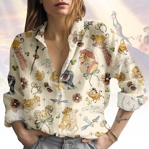 Puede incluir: Camisa crema con botones con un patrón repetido de personajes e imágenes de El Rey León. El diseño incluye a Simba, Timón, Pumba y Zazú. La frase "It means no worries" también es visible.