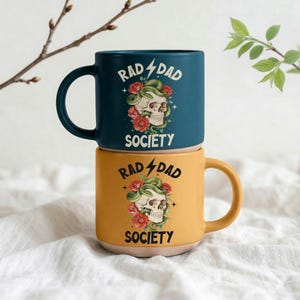 Könnte beinhalten: Zwei Keramikbecher übereinander gestapelt. Der obere Becher ist marineblau, der untere senfgelb. Jeder Becher zeigt ein Totenkopf-Design mit einer Schlange, Rosen und den Worten "RAD DAD SOCIETY".