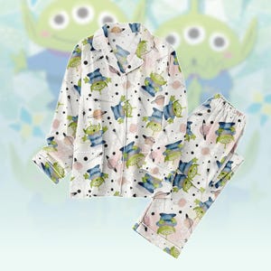 Könnte beinhalten: Weißes Pyjama-Set mit einem sich wiederholenden Muster aus grünen, blauen und rosa Alien-Charakteren, Planeten und Sternen. Das langärmlige Oberteil hat eine Knopfleiste und einen Reverskragen. Die Pyjamahose hat einen elastischen Bund.