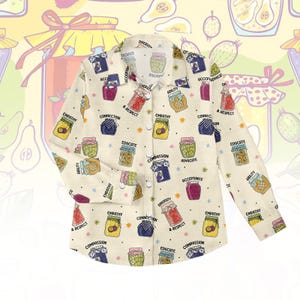 Può includere: Camicia a maniche lunghe color crema con un motivo di barattoli di marmellata colorati e parole come "Empatia", "Educare" e "Diversità". La camicia ha un colletto e polsini abbottonati. Misure in cm.