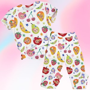 Könnte beinhalten: Weißes Pyjama-Set mit einem farbenfrohen Fruchtdesign. Das Set besteht aus einem kurzärmeligen Oberteil und einer Hose, die mit Illustrationen von lächelnden Früchten wie Äpfeln, Bananen und Erdbeeren sowie blauen Sternen bedeckt sind.