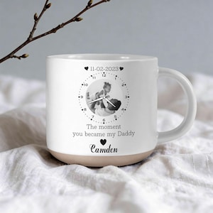 Könnte beinhalten: Eine weiße Keramik-Tasse mit schwarzem Henkel und dem Text "But First, Coffee" in schwarzer, handschriftlicher Schrift. Die Tasse steht auf einer weißen Oberfläche.