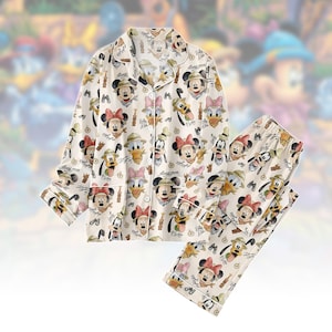 Op de afbeelding: Crèmekleurige pyjamaset met een herhaald patroon van stripfiguren, waaronder Mickey en Minnie Mouse, Donald Duck en Pluto. De top met lange mouwen heeft een knoopsluiting en witte bies. De bijpassende broek heeft een elastische tailleband.