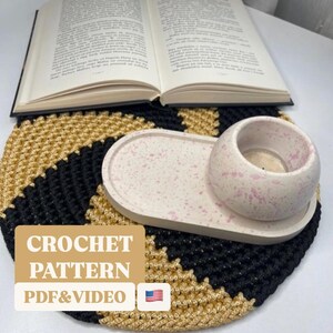 May include: Crochet pattern for a black and gold round coaster with a white speckled oval tray and a white speckled candle holder. The text "CROCHET PATTERN PDF & VIDEO" is on the coaster. An American flag is in the lower right corner.
