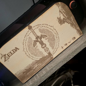 Peut inclure: Une protection en bois clair pour console de jeu portable avec une gravure laser représentant l'épée de légende de Zelda, entourée de motifs circulaires, de texte Hylien et d'une petite silhouette grimpant sur une falaise.