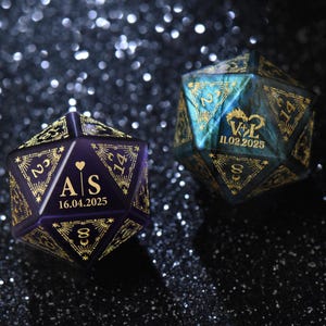 May include: Two personalized, multi-sided dice. One is purple with gold text, displaying initials and a date. The other is teal and gold, also with initials and a date. Both dice are for a wedding.