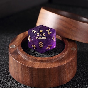 May include: A purple, personalized D20 die with gold numbering and the initials "S&K" and the date "20.05.2025" engraved on it. The die rests in a small, open wooden box with a dark interior.
