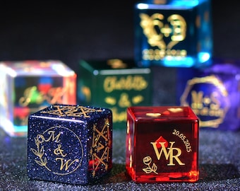 May include: A collection of personalized dice in various colors, including blue, red, green, and purple. Each die features custom gold lettering and designs, with some displaying initials, dates, and floral accents. The dice are arranged on a dark, glittery surface.