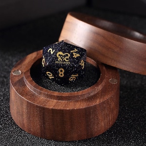 May include: A dark blue, twenty-sided die with gold numbering and a personalized inscription, resting in a wooden box. The die has a speckled appearance and is set against a black interior. The wooden box is open, revealing the die.