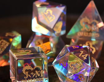 May include: A set of iridescent, transparent polyhedral dice featuring gold-engraved dragon silhouettes, the initials J+M, and the date 09.13.2025. The dice have sharp edges and display a multi-colored, reflective sheen across their surfaces.