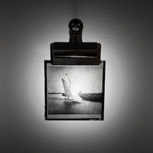 May include: Black and white photograph of a sailboat on the water, held by a metal clip. The image is a vintage print, showing the boat with its sails up, moving across the water. The clip is attached to a wall.
