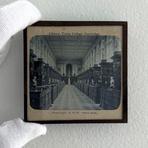 以下が含まれることがあります： 本棚と胸像が並ぶ図書館の内部の白黒写真。上部には「Library, Trinity College, Cambridge」の文字が見えます。画像は濃い茶色のフレームに入っています。