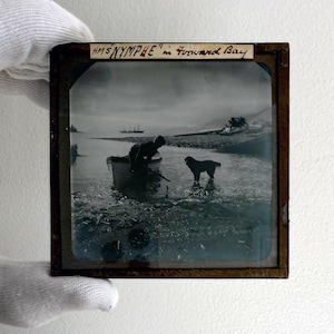 May include: Antique photograph in a wooden frame depicting a scene with a boat, a person, and a dog in the water. The text "HMS NYMPHE" in Forward Bay" is written at the top. The image has a vintage aesthetic.