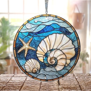 Peut inclure: Vitrail rond représentant une coquille, une étoile de mer et des vagues bleues, blanches et beiges. L'œuvre d'art est encadrée d'une bordure dorée et suspendue à une chaîne argentée. Une pièce décorative pour un thème côtier, environ 20 cm de diamètre.