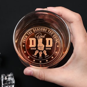 May include: A clear, round whiskey glass with the words "FROM THE REASONS YOU DRINK" and "Best DAD EVER" etched on the front. The design includes a fist and three smaller fists, with names below. The glass is held by a hand.