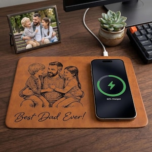 May include: A brown wireless charging pad with a family portrait and the text "Best Dad Ever!". A smartphone is charging on the pad, displaying a green charging symbol. A framed photo and a succulent plant are also visible.