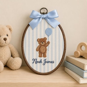 May include: An oval embroidery hoop featuring a light blue and white striped fabric background. The centre displays a brown teddy bear holding a blue balloon, with the name Noah James embroidered in dark blue script below the bear.
