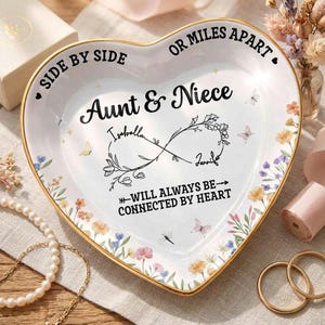 May include: Heart-shaped ceramic dish with gold trim, featuring floral and butterfly designs. Text reads "Aunt & Niece" with names and the phrase "Will Always Be Connected By Heart". Decorative item.