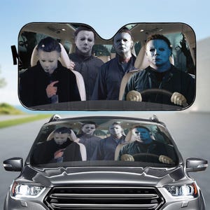 Puede incluir: Parasol de coche negro con un gráfico de Michael Myers de la serie de películas de terror Halloween. La imagen muestra a Myers en un coche con otros personajes. El parasol está diseñado para un parabrisas de coche.
