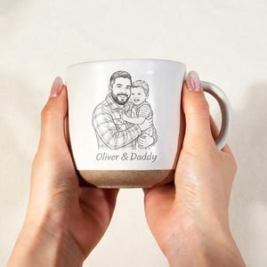 May include: White ceramic mug featuring a sketch of a father and child, with the text "Oliver & Daddy". The mug has a brown base and a curved handle. The image is held by a person.