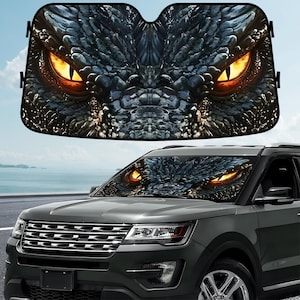 May include: A black car sunshade featuring a close-up of a dragon's face with glowing orange eyes. The dragon's scales are a mix of dark blue and gray, creating a textured appearance. The sunshade is installed on the windshield of a dark gray SUV.