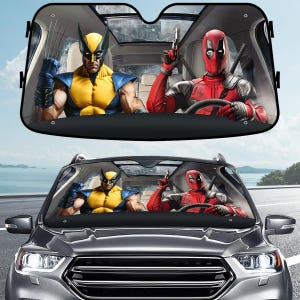 May include: Car sunshade featuring Wolverine in a yellow and blue costume and Deadpool in a red suit. The characters are depicted inside a car, with Wolverine in the passenger seat and Deadpool driving. The sunshade is black.