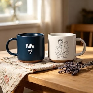 May include: Two ceramic mugs on a wooden table. One is navy blue with "PAPA" and a heart. The other is white with a sketch of a man and child, with the text "Leo & Papa". Both mugs have a brown base.