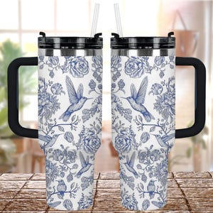 Puede incluir: Dos tazas de viaje de acero inoxidable con asas y tapas negras. Las tazas presentan un fondo blanco con un diseño floral y colibríes azules. Cada taza tiene una pajita transparente.
