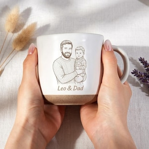 May include: A white ceramic mug with a brown base, held by two hands. The mug features a black and white illustration of a man holding a child, with the text "Leo & Dad" below the illustration. The mug has a curved handle.