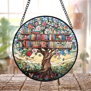 May include: Round stained glass window art featuring a tree with bookshelves for branches. The tree is filled with colorful books, flowers, and butterflies. The window is framed in black and hangs from a chain.
