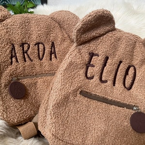 Könnte beinhalten: Zwei braune Rucksäcke in Teddybärform mit den gestickten Namen "ARDA" und "ELIO". Jeder Rucksack hat eine Reißverschluss-Vordertasche, Bärenohren und einen runden, braunen Anhänger. Die Rucksäcke sind aus weichem, strukturiertem Material.