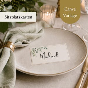 May include: A rectangular white place card with a green leaf illustration and the name Michael in black script, resting on a beige ceramic plate next to a sage green linen napkin held by a gold ring, with text reading Sitzplatzkarten and Canva Vorlage.