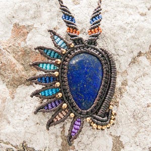 May include: A dark brown macrame necklace featuring a large, deep blue lapis lazuli gemstone pendant. The pendant is surrounded by an intricate, multi-coloured macrame design with gold beads.
