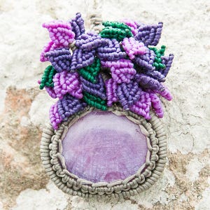 May include: A handcrafted macrame pendant with a large, oval, lavender-coloured stone. The stone is framed by a grey macrame border. Above the stone, there is a cluster of purple, green, and pink macrame leaves.