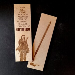 May include: Two beige bookmarks with laser-etched designs. One features text that reads "JOHN WILL COME FOR YOU AND YOU WILL DO NOTHING... BECAUSE YOU CAN DO NOTHING" and an image of a man holding a gun. The other bookmark has a pencil design.