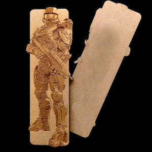 Puede incluir: Un marcapáginas de madera con una imagen grabada con láser de un personaje de videojuego con un traje futurista que sostiene un arma. El personaje está de pie con las piernas ligeramente separadas y los brazos cruzados. El marcapáginas es de color marrón claro y tiene un acabado liso y pulido.