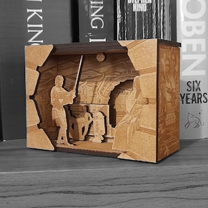 May include: A wooden box containing a diorama of a Star Wars scene. The scene features a character holding a lightsaber, another character seated, and a droid. The box is brown with a black base. The box has a label on the side that reads "Star Wars" and "Six Years".