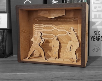 Puede incluir: Una caja de sombra de madera que representa una escena de la película 'Karate Kid'. La caja de sombra presenta un cuadro de torneo de karate con el texto 'All Valley Karate Championship' y tres figuras de luchadores de karate.