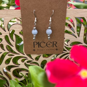 May include: A pair of silver earrings with light blue round gemstone beads. The earrings are displayed on a brown card with the text "Picer Design Studio".
